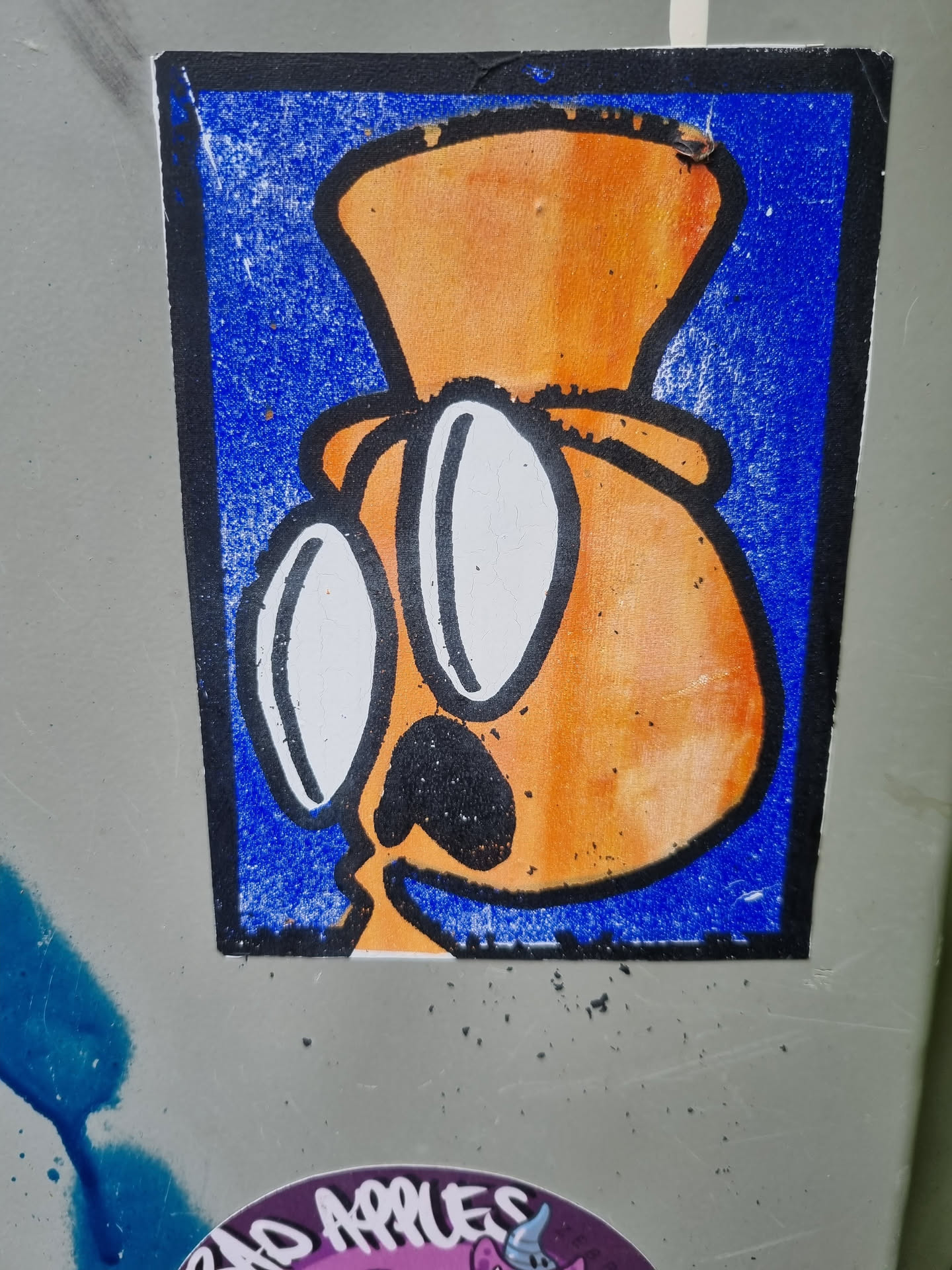A sticker featuring an orange cartoon character wearing a hat and glasses. The character has a simple design with a black nose and mouth. The background of the sticker is blue. 