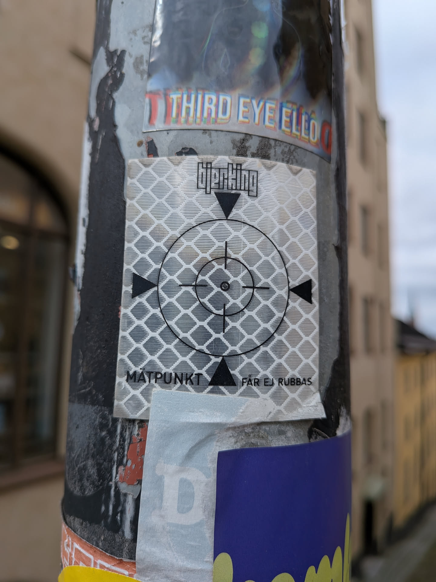 The sticker is predominantly silver and white with a reflective, diamond-patterned background. It features a black targeting reticle in the center, consisting of a circle with crosshairs, a smaller circle in the middle, and four triangular markers at the cardinal points. Above the reticle, there is black text that appears to read 'Hjerkking' in a stylized font. Below the reticle, the words 'MÄTPUNKT' are visible on the left, and 'FÅR EJ RUBBAS' is visible on the right, both in black, uppercase lettering. The sticker is attached to a dark, weathered surface, likely a utility pole, and is partially overlapped by other stickers.