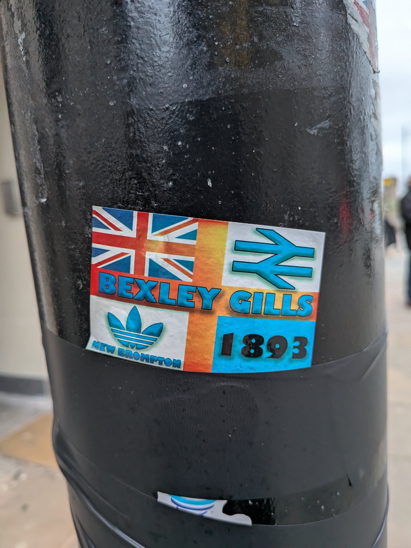 A rectangular sticker adhered to a textured black pole. The design is a collage of elements on a background that transitions from orange to yellow to blue. The top-left section features the Union Jack flag. Below it, on the bottom-left, is a blue Adidas trefoil logo with the text 'NEW BROMPTON' in smaller blue letters underneath. The top-right section displays a blue railway symbol (double arrow). Prominently in the center, in bold blue letters, is 'BEXLEY GILLS'. The bottom-right corner shows the numbers '1893' in large blue font. The sticker shows some signs of wear, with slight peeling at the edges.
