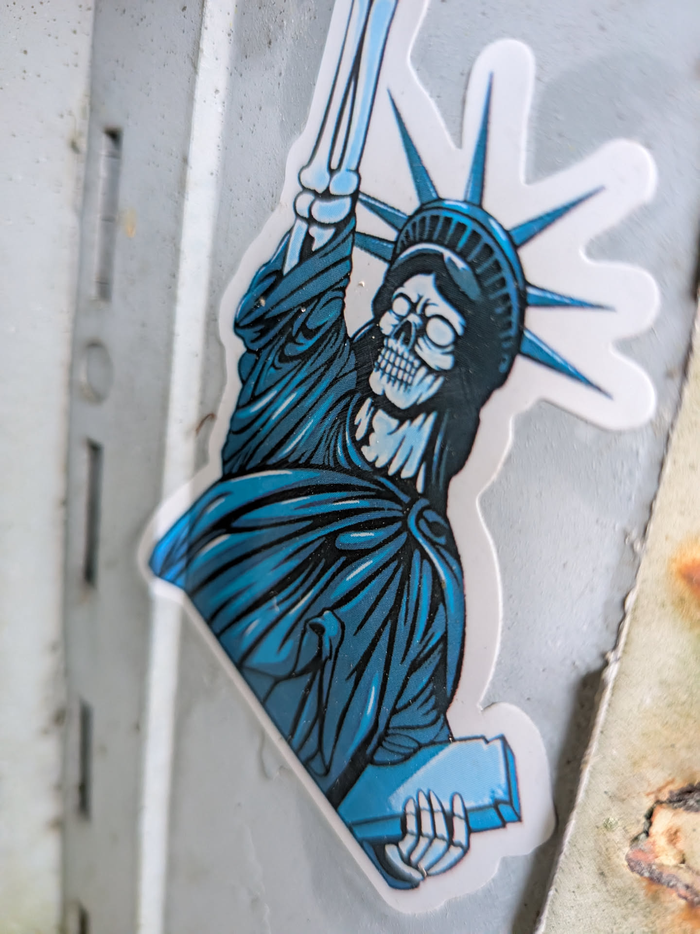 A die-cut sticker featuring the upper torso of the Statue of Liberty reimagined as a skeletal figure. The figure wears the classic seven-point spiked crown and draped robe in vibrant shades of blue, teal, and deep black shading. Its face is an exposed white skull with hollow eye sockets and a grim toothy grin, accompanied by a bony neck and skeletal hand holding the tablet below. An arm composed of bare arm bones raises the torch towards the top of the design, all enclosed by a white border.