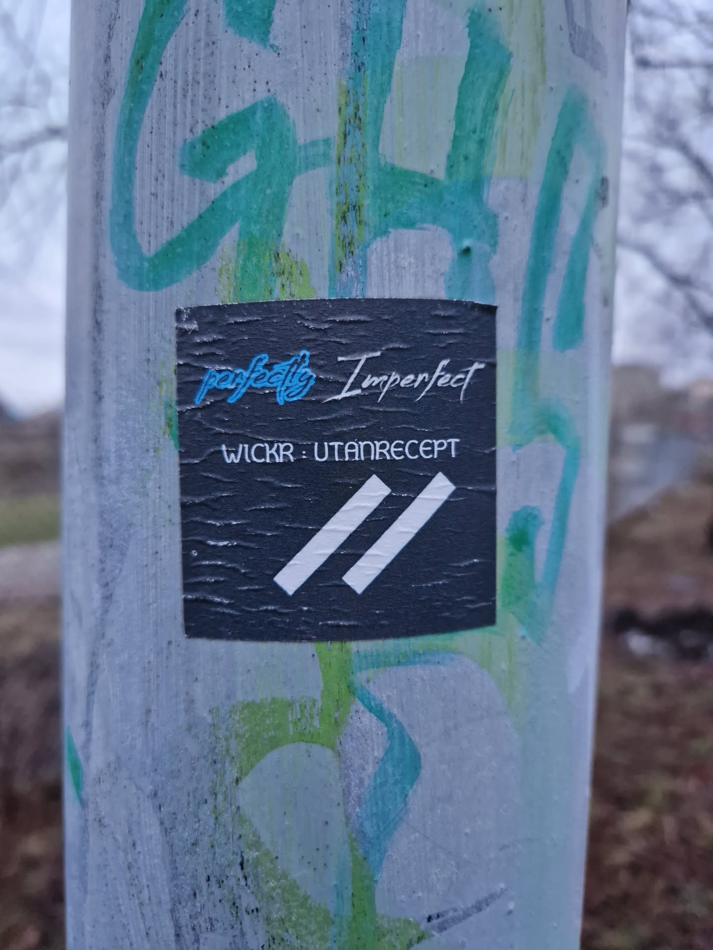 A black sticker with the text "perfectly Imperfect" and "WICKR: UTANRECEPT" and a white double parallel lines graphic. The sticker is attached to a pole with other graffiti on it.