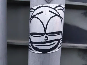 A black and white sticker featuring a cartoon face with a slight smile. The sticker is affixed to a weathered metal pole, giving it a slightly distressed appearance.  The style is simplistic, yet expressive. 