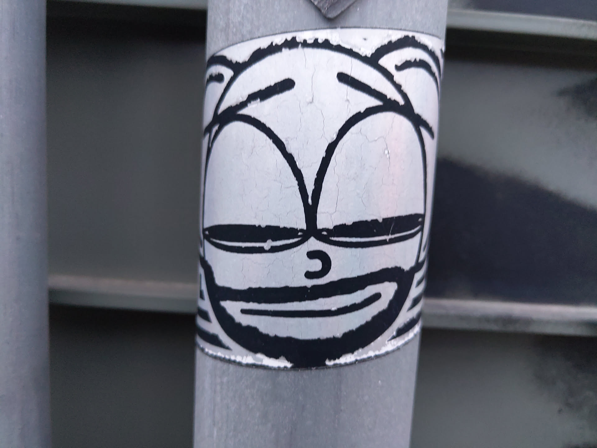 A black and white sticker featuring a cartoon face with a slight smile. The sticker is affixed to a weathered metal pole, giving it a slightly distressed appearance.  The style is simplistic, yet expressive. 