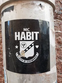 Black and white sticker with a logo featuring the text "MC HABIT" and a crest. The crest includes a star, a symbol resembling a stylized yin-yang, a diamond pattern, and a heart.  The text "MASTER OF CEREMONIES" is written in an arc below the crest.