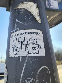 This weathered sticker on a gray pole depicts a black and white cartoon. On the left, a drum with sticks and the letters "DD". On the right, a bear-like figure with "DD" on its stomach, is holding up signs. The sticker has the text "@DIFFERENTDRUMBLN" at the top.
