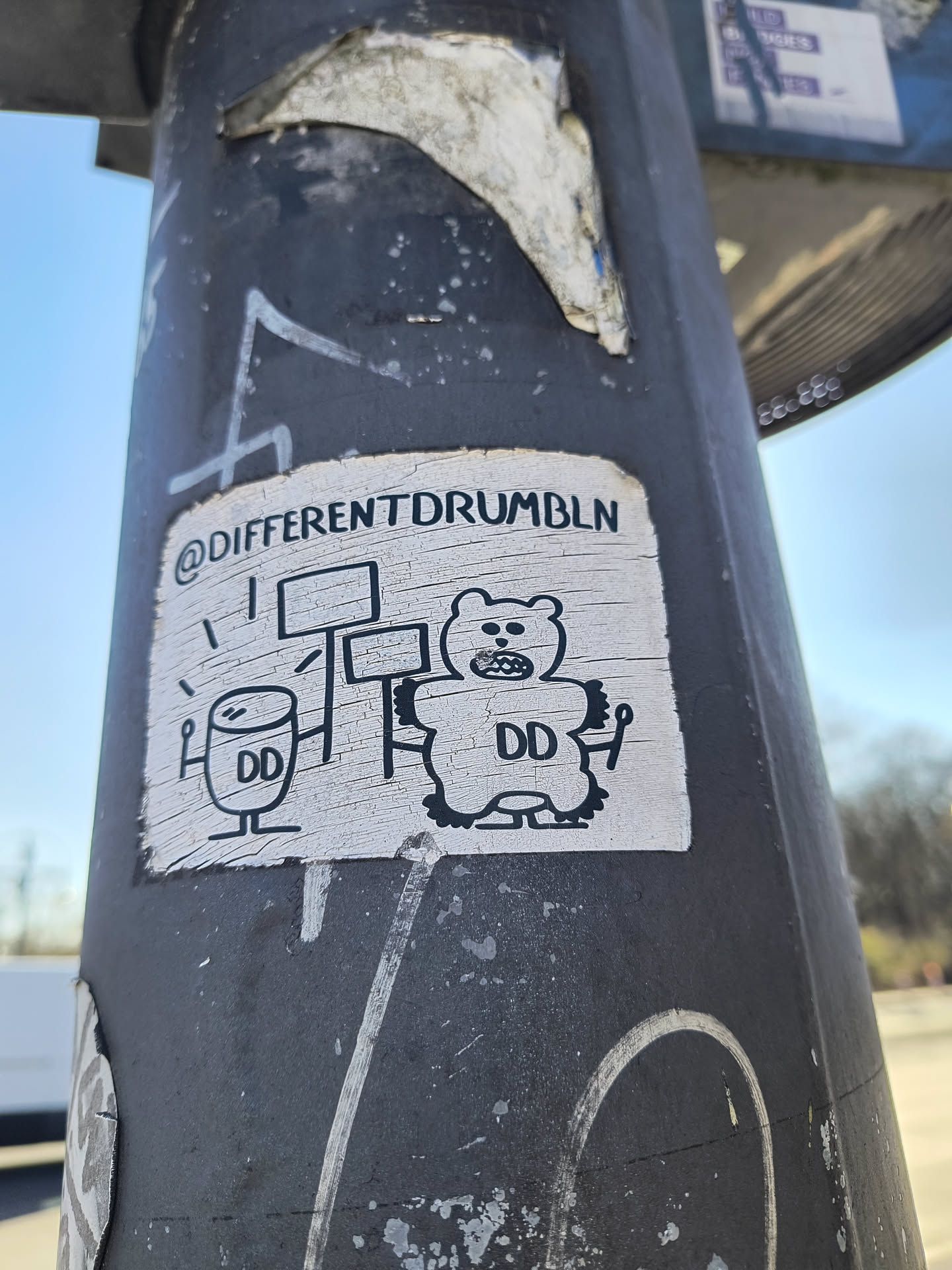 This weathered sticker on a gray pole depicts a black and white cartoon. On the left, a drum with sticks and the letters "DD". On the right, a bear-like figure with "DD" on its stomach, is holding up signs. The sticker has the text "@DIFFERENTDRUMBLN" at the top.