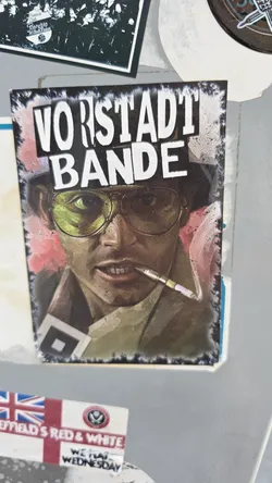 Street sticker A vertical rectangular sticker depicting an artistic portrait of Raoul Duke from Fear and Loathing in Las Vegas, wearing yellow-tinted aviator glasses, a bucket hat, and holding a cigarette holder between his teeth. Above the character, the bold white text 'VORSTADT BANDE' is displayed against a dark smoky background with distressed, burned-style borders.