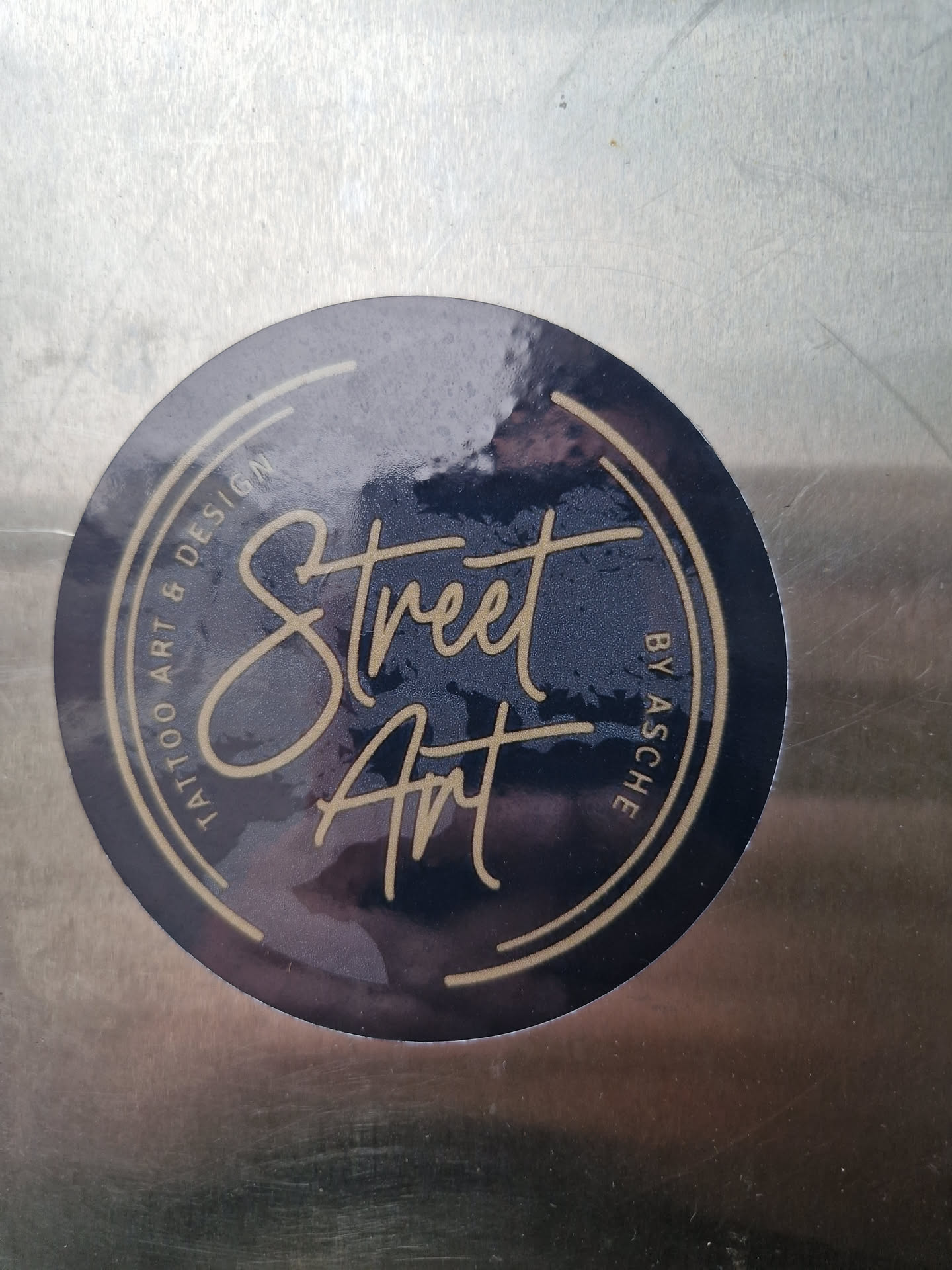 Round sticker with a dark blue background and gold circular border. The text "Street Art" is written in a stylized script font in the center.  The smaller text around the circle says "Tattoo Art & Design by Asche".