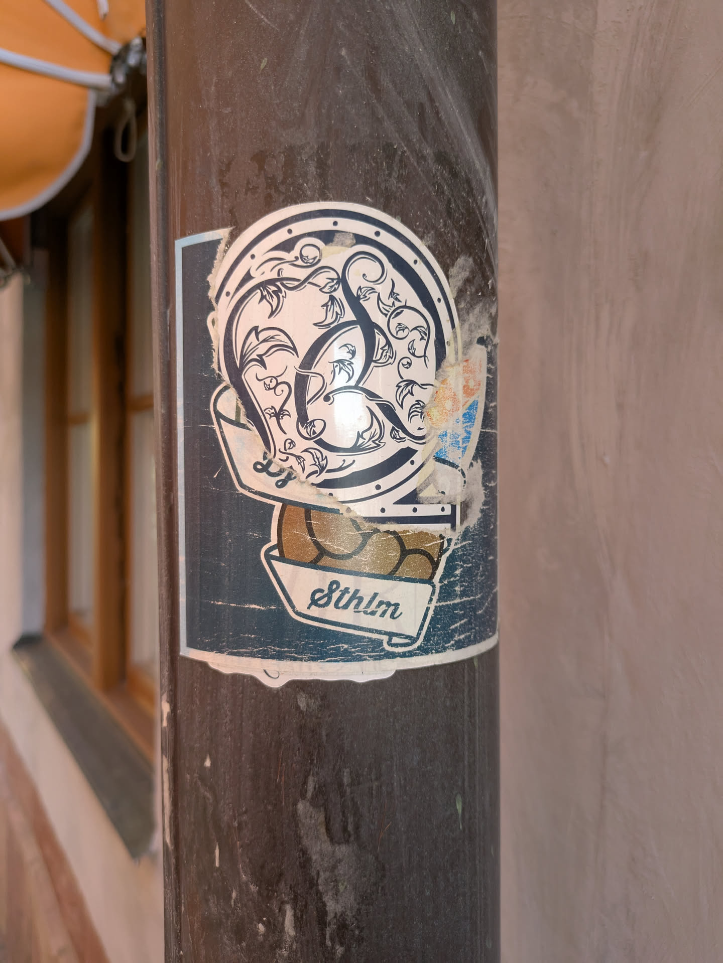 A weathered sticker on a pole featuring an ornate, circular design above a container labeled 'Sthlm'. The top part of the sticker has an intricate pattern resembling intertwined vines or floral elements, all enclosed within a circle. Below this design is a container filled with what appears to be golden nuggets or similar round objects. The sticker has a blue and white color scheme with a slightly distressed appearance, suggesting it has been exposed to the elements for some time.