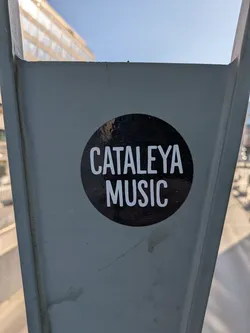 This is a round sticker featuring the text "CATALEYA MUSIC" in white letters on a black background. The sticker is affixed to a grey metal structure, possibly part of a bridge or architectural element. The lighting is bright, suggesting an outdoor setting.
