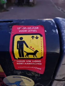 A red and yellow sticker warning about the danger of injury and asking to keep dogs close on a leash.  The sticker contains a pictogram of a person walking a dog.