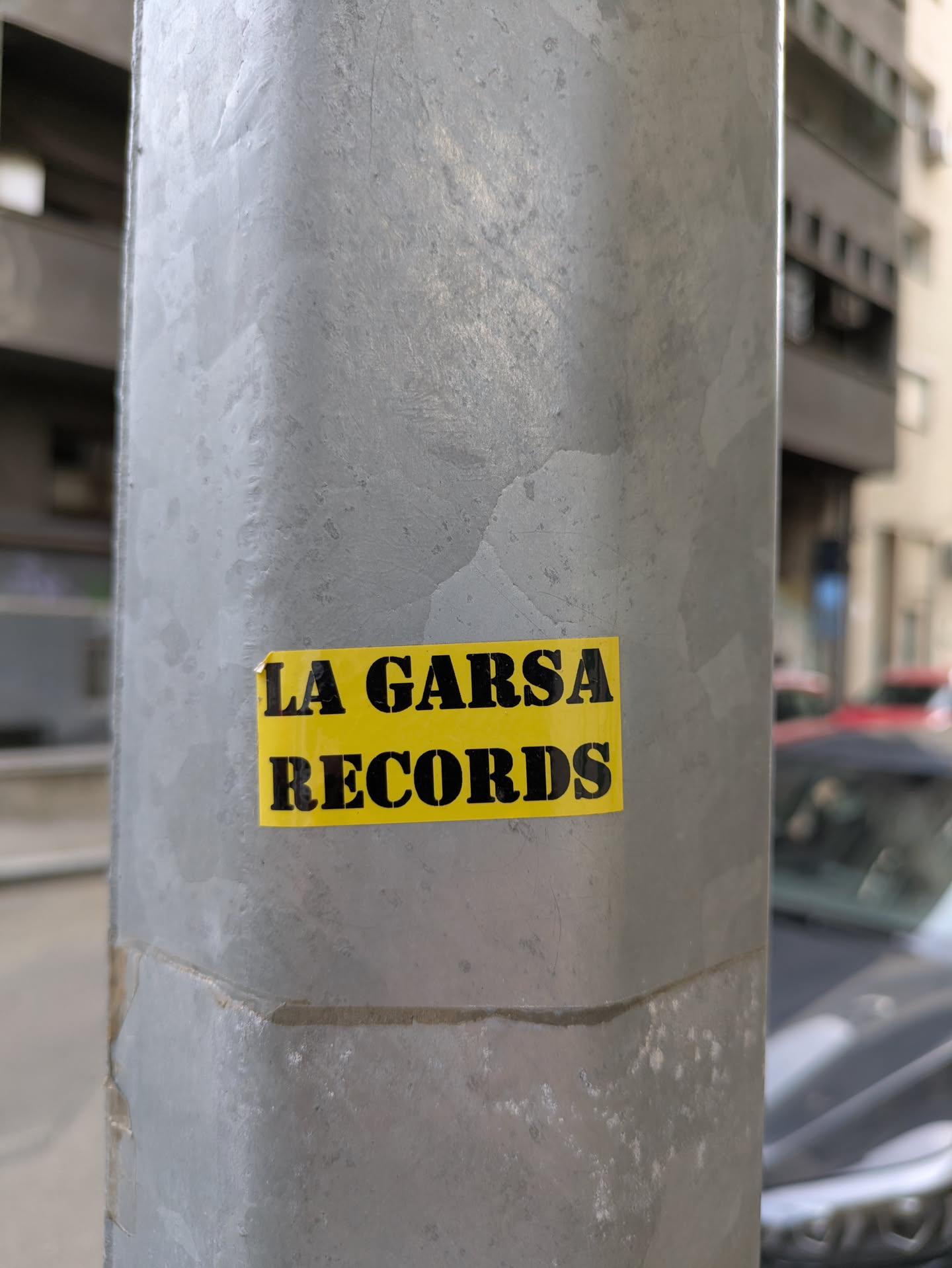 A rectangular yellow sticker with black lettering on a gray metal pole. The sticker reads "LA GARSA RECORDS". The pole is weathered and shows signs of wear and tear.