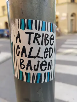 A sticker with a simple design featuring the text "A Tribe Called Bajen" written in black marker over a white background. The sticker has blue and black stripes on its top and bottom. It is affixed to a grey pole.