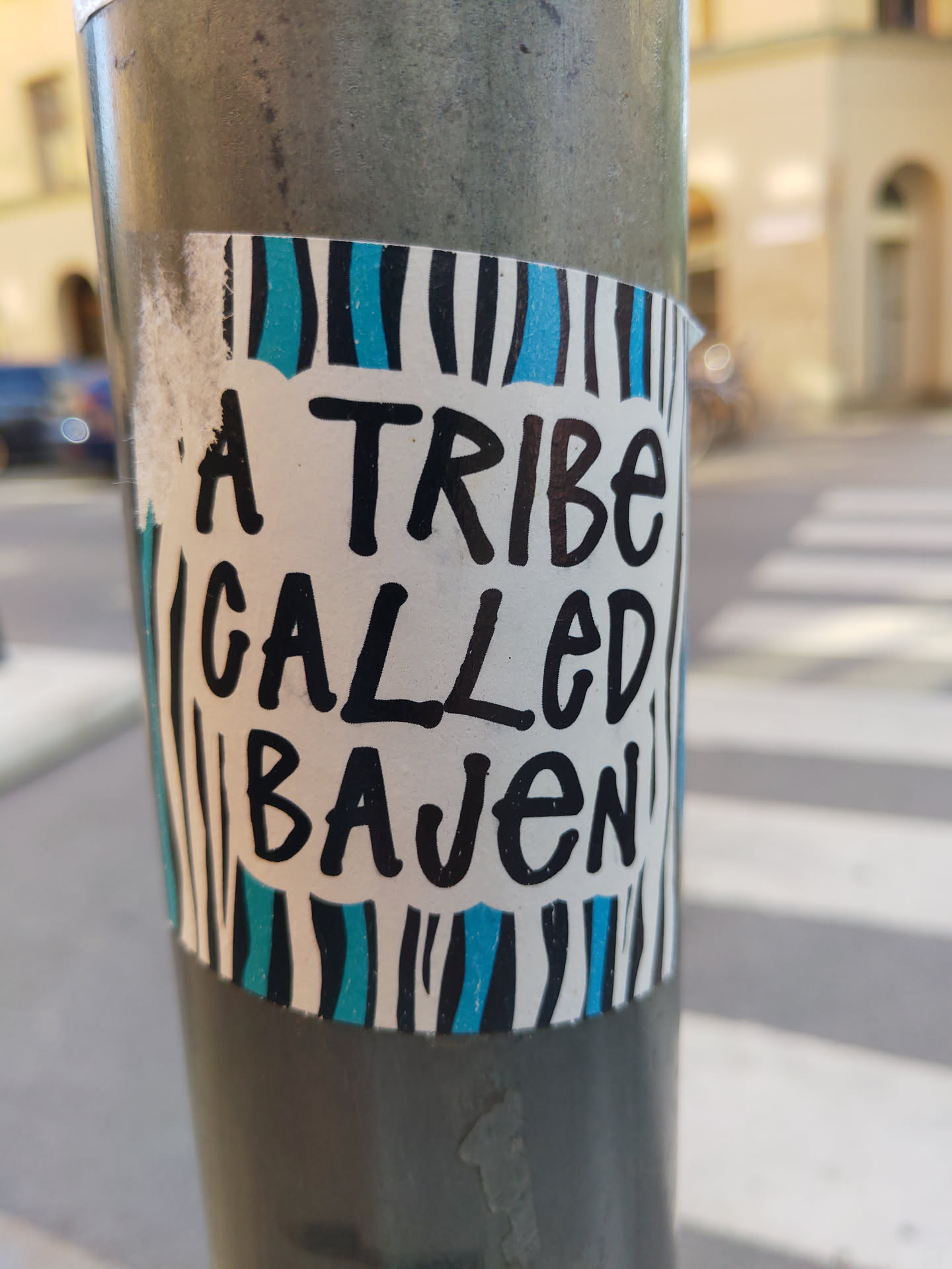 A sticker with a simple design featuring the text "A Tribe Called Bajen" written in black marker over a white background. The sticker has blue and black stripes on its top and bottom. It is affixed to a grey pole.