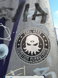 Round black and white sticker with a central image of a stylized face with an eye in the center and tentacles. The text around says "Question Everything", and it's also signed by Carl Kenz. The sticker has a vintage or grunge aesthetic.