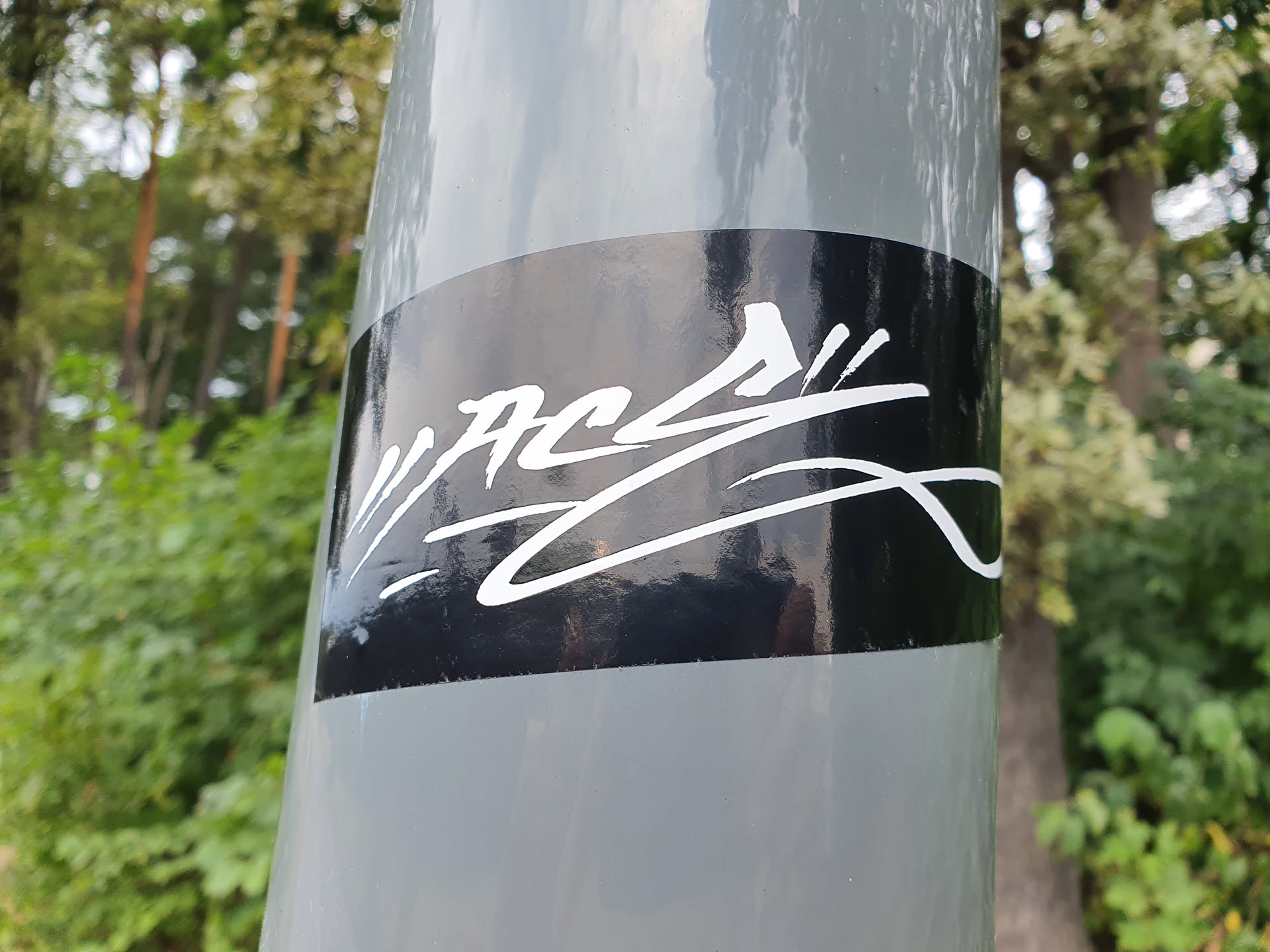 A black and white sticker with a graffiti-style design on a gray pole. The sticker has a glossy finish and the design is in white against a black background. The background shows green vegetation, indicating an outdoor setting.
