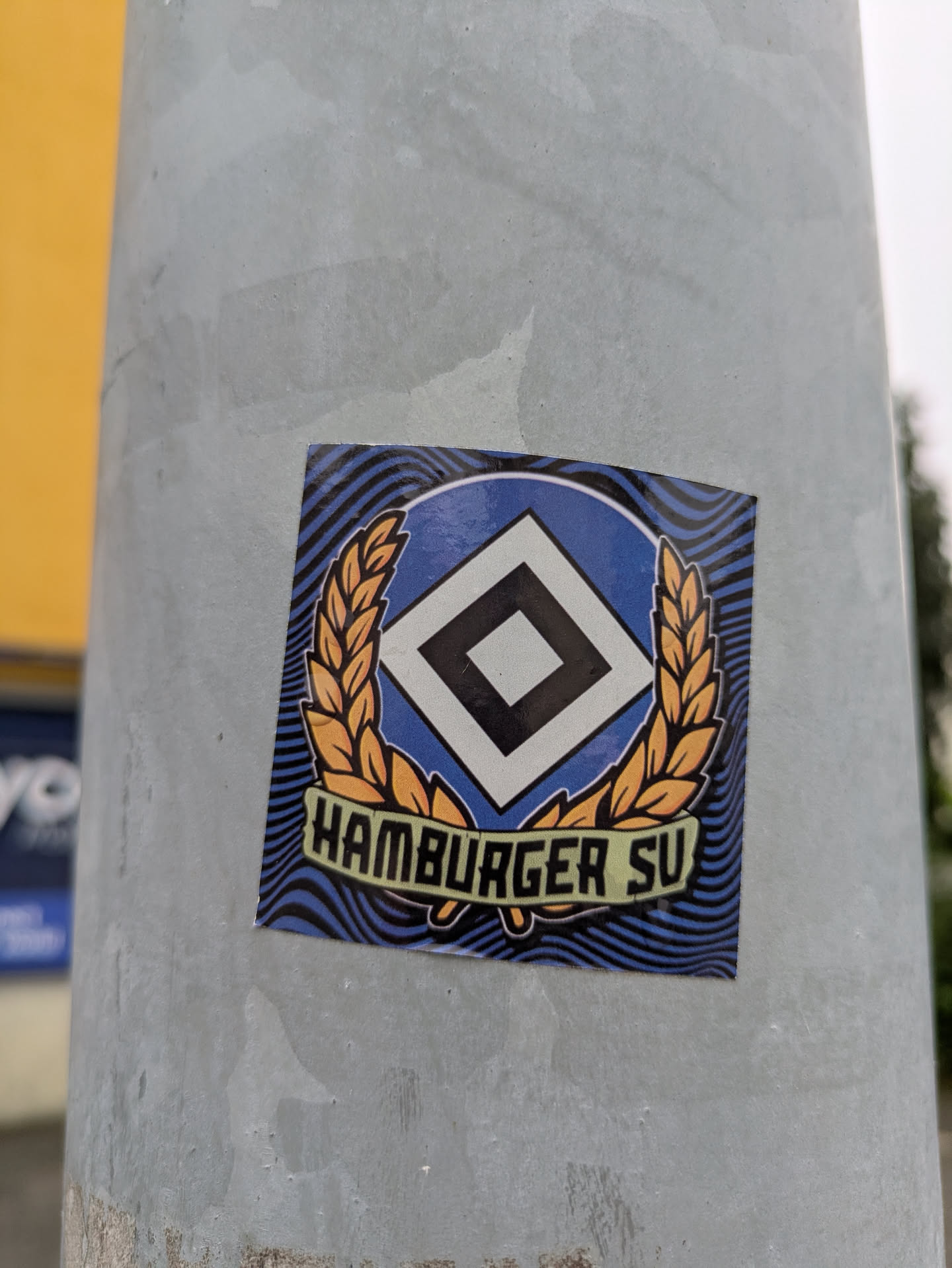 The sticker features the crest of Hamburger SV. The central design is an oval with a blue background, containing a white diamond outlined in black, which in turn frames a smaller black diamond. This oval is flanked by two golden-orange laurel wreaths. Below the main emblem, a light green banner displays the text 'HAMBURGER SV' in black capital letters. The background of the sticker, outside the crest, is a dynamic pattern of wavy black lines on a dark blue base. The sticker is square with slightly rounded corners and is affixed to a grey metal pole.