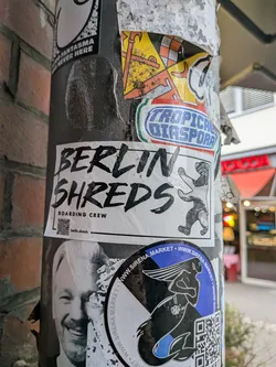 Street sticker A rectangular white sticker adhered to a metal post. It features bold, hand-drawn brush lettering reading 'BERLIN SHREDS' across two lines, with 'BOARDING CREW' printed in a cleaner sans-serif font underneath. To the right is an illustration of the iconic Berlin bear riding a skateboard. At the bottom left, there is a small QR code next to an Instagram handle: 'berlin.shreds'.