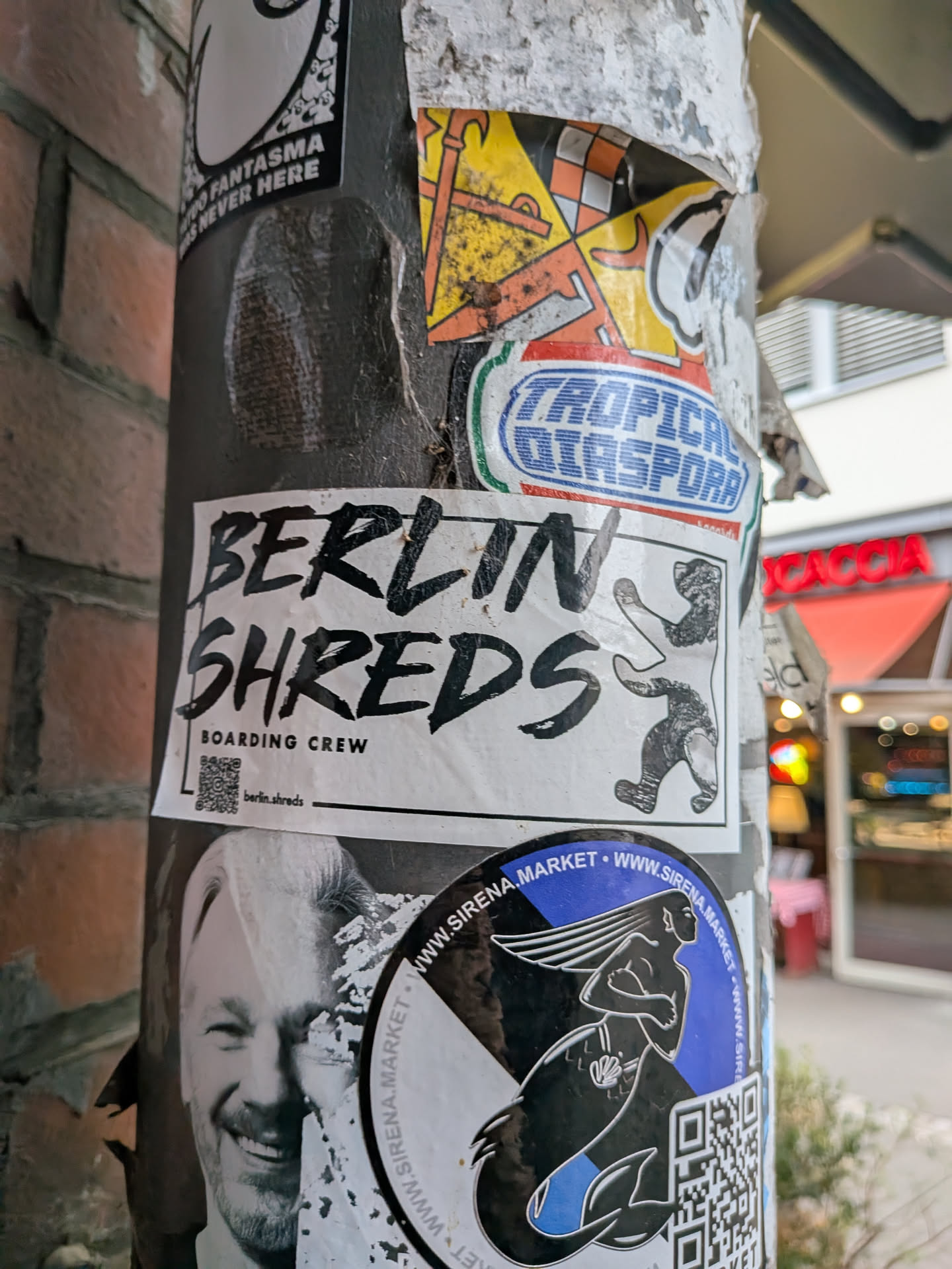 A rectangular white sticker adhered to a metal post. It features bold, hand-drawn brush lettering reading 'BERLIN SHREDS' across two lines, with 'BOARDING CREW' printed in a cleaner sans-serif font underneath. To the right is an illustration of the iconic Berlin bear riding a skateboard. At the bottom left, there is a small QR code next to an Instagram handle: 'berlin.shreds'.