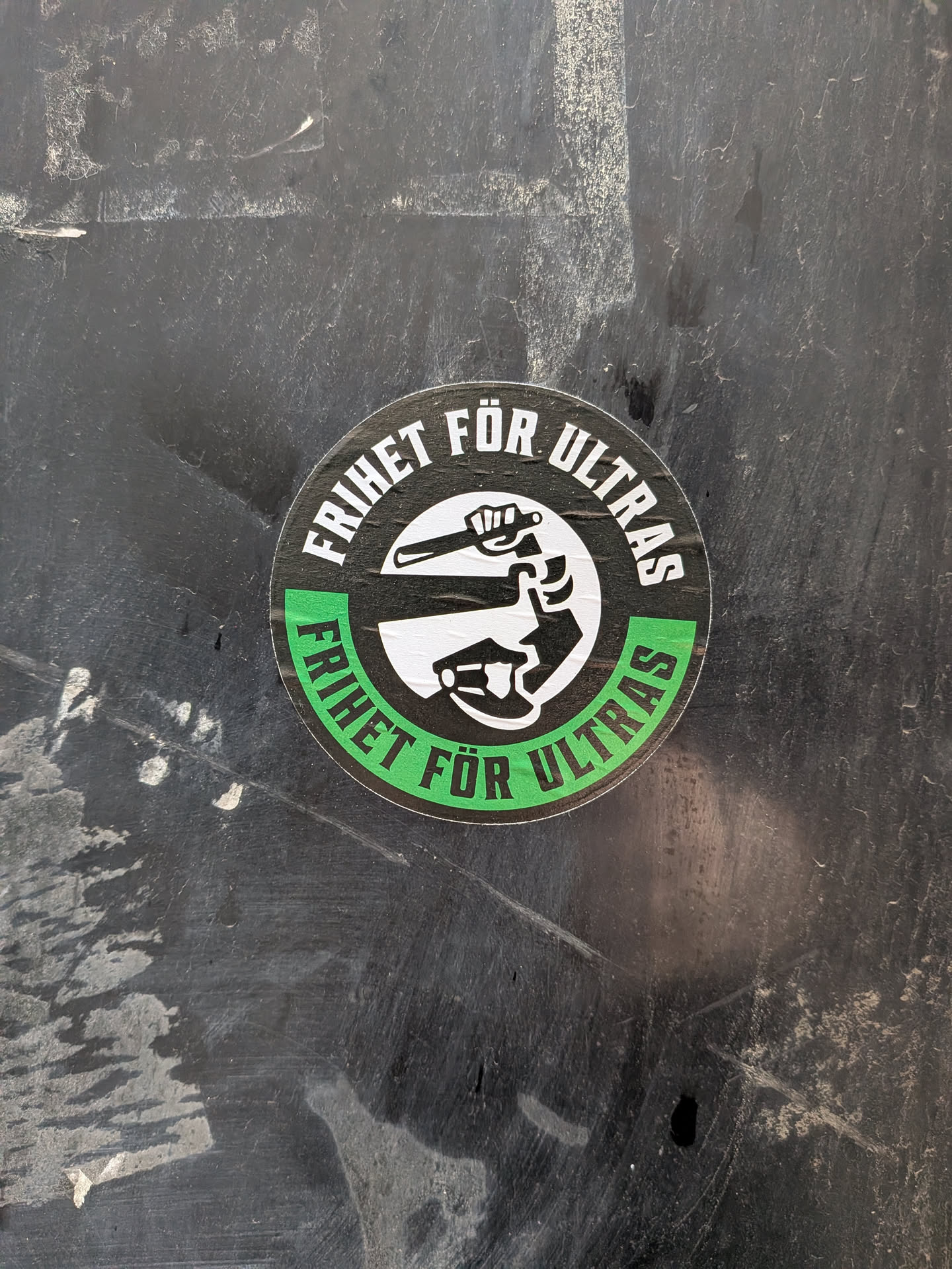 The sticker is circular with a black border. The top half of the border contains white text that reads 'FRIHET FÖR ULTRAS' in a bold, sans-serif font. Inside this text, there is a white circle containing a black silhouette of a clenched fist holding a baton or club. The bottom half of the border is a vibrant green, also containing white text that reads 'FRIHET FÖR ULTRAS' in the same font. The overall design is stark and impactful, using a limited color palette of black, white, and green.