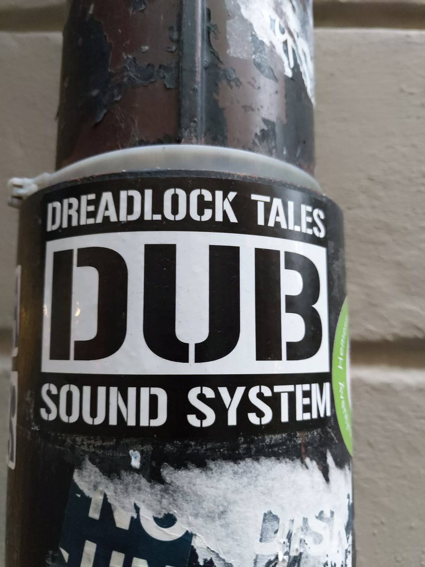 A black and white sticker with the text "Dreadlock Tales" at the top and "DUB Sound System" at the bottom, with the word "DUB" prominently displayed in a large, bold font in the center. The sticker is affixed to a weathered, dark-colored metal surface.