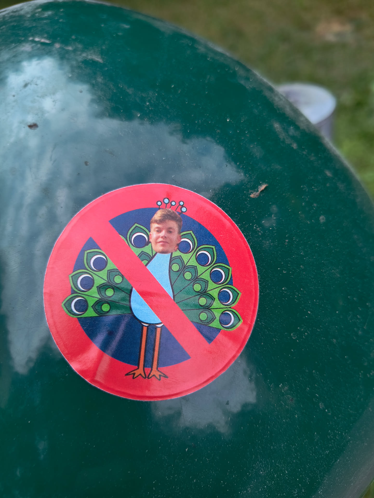 A round sticker with a red circle and slash over a cartoon peacock.  The peacock has an unusual head; a person's face is depicted in place of the peacock's head. The sticker is affixed to a dark green surface. 
