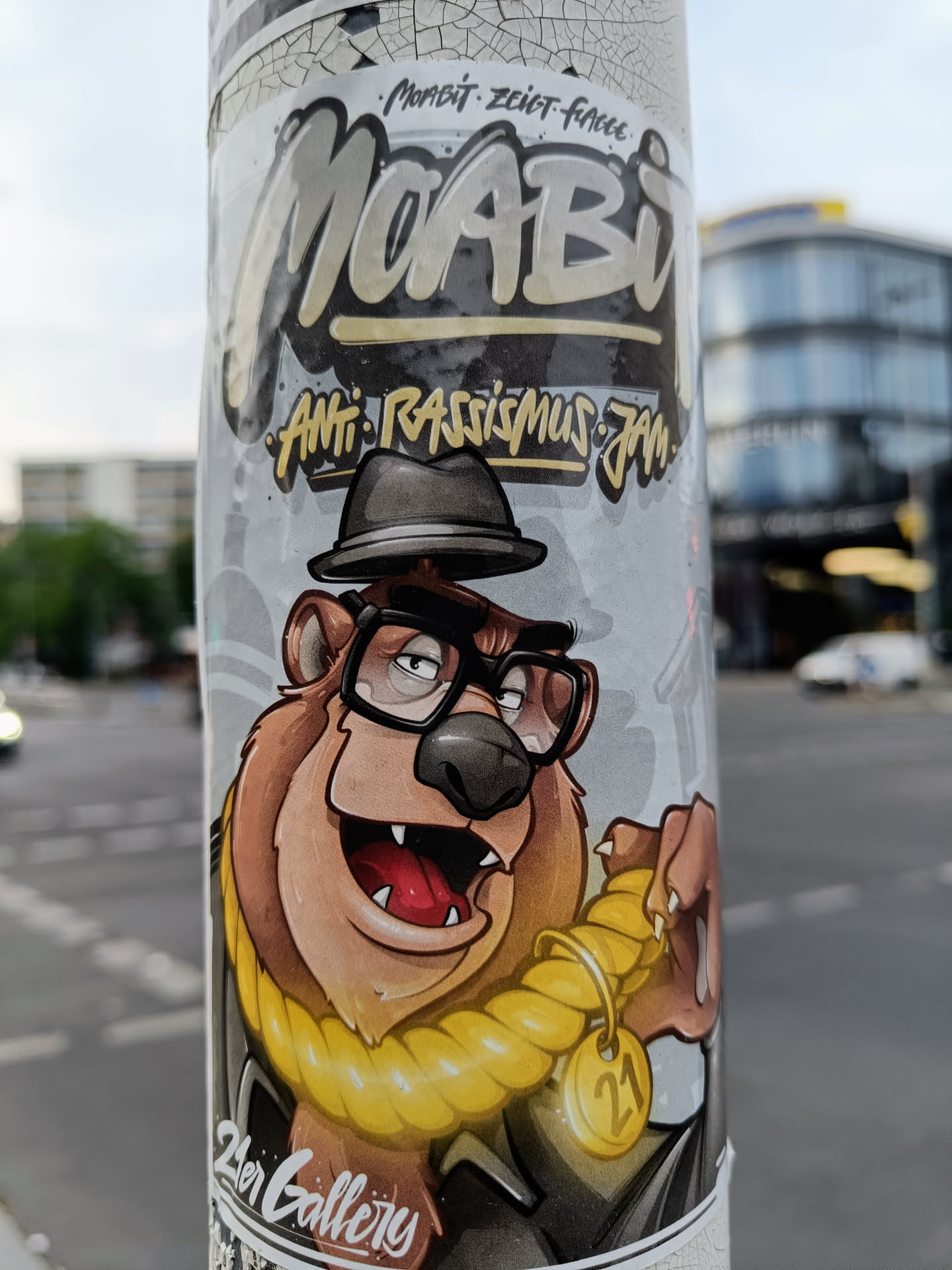 Sticker featuring a cartoon illustration of a bear wearing a hat and glasses, with a gold rope around its neck and a tag with the number 21.  The sticker has the words "Moabit", "Anti- Rassismus jetzt", and "2er Gallery" on it.
