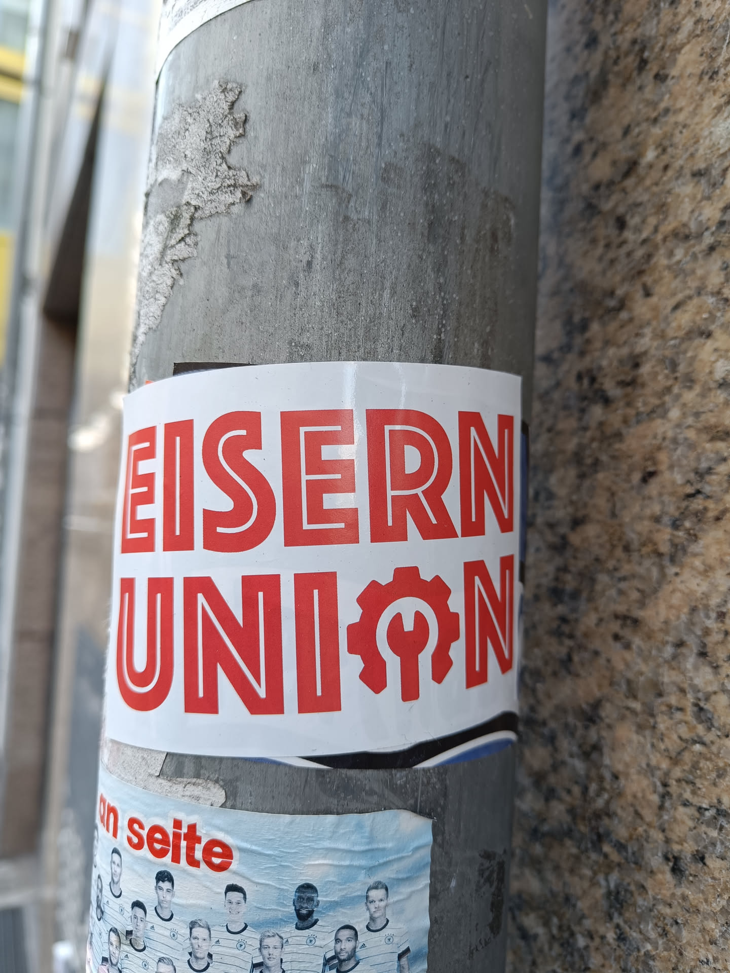 A sticker featuring the text "EISERNE UNION" in bold red letters, accompanied by a red logo that seems to depict a gear with a wrench inside. The sticker is affixed to a gray metal pole.