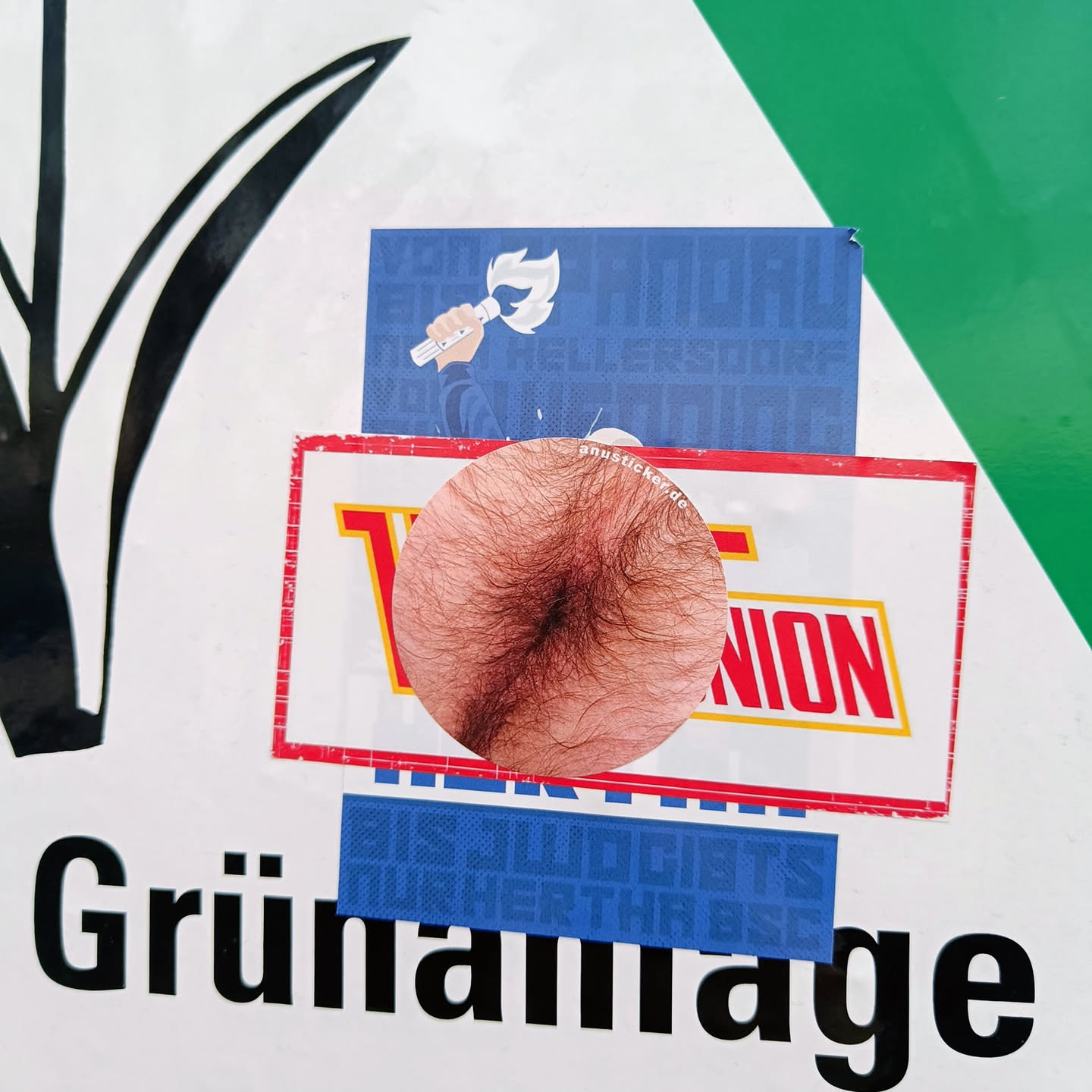 A round sticker depicting a hairy belly button is affixed to a sign.  Other stickers are layered on top and underneath it. The main sign is predominantly white with the word "Grünanlage" (German for green space or park) in bold black lettering.