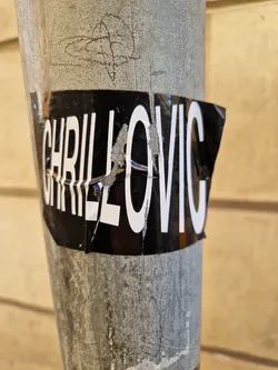 A black and white sticker with the text "CHRILLOVIC" in bold, sans-serif font. The sticker is slightly damaged and torn at the edges, adding a bit of urban grit to its aesthetic. It's affixed to a weathered gray metal pole, against a backdrop of light beige wood siding.