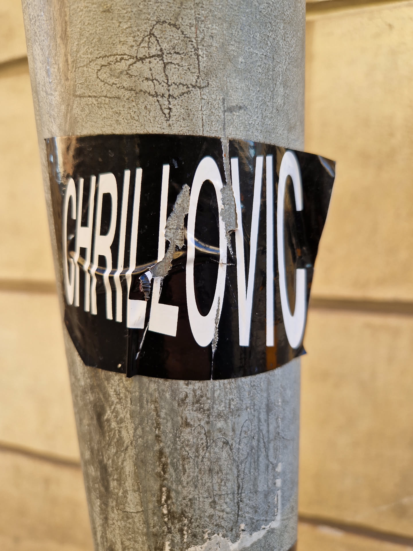 A black and white sticker with the text "CHRILLOVIC" in bold, sans-serif font. The sticker is slightly damaged and torn at the edges, adding a bit of urban grit to its aesthetic. It's affixed to a weathered gray metal pole, against a backdrop of light beige wood siding.