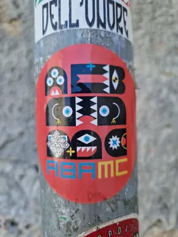Round sticker with a predominantly red background. It features a stylized face design composed of geometric shapes and patterns in black, white, and teal. The design incorporates various symbols, including eyes, triangles, and crosses. At the bottom, the letters "ABAMC" are written in a colorful, stylized font.
