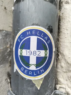 The sticker is circular with a blue outer ring, followed by a yellow inner ring. The central design features a white cross on a blue background, reminiscent of the Greek flag. On either side of the cross, there are green laurel branches. The year '1982' is prominently displayed in white digits within the center of the cross. The text 'FC HELLAS' curves along the top inner edge of the blue ring, and 'BERLIN' curves along the bottom inner edge, both in white capital letters.