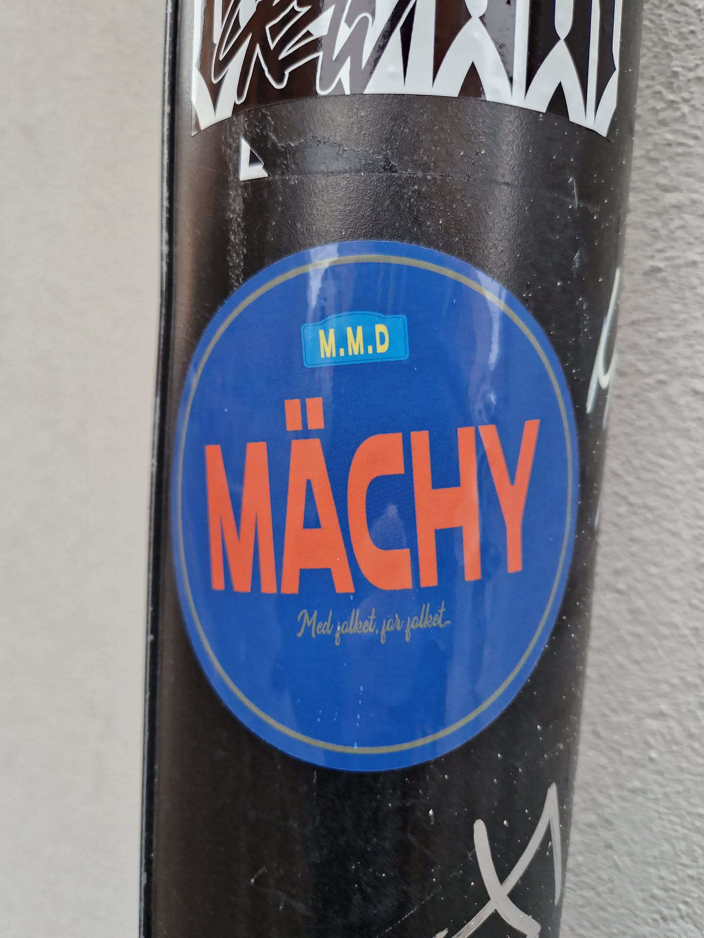 A round blue sticker with the word "Mächy" in large orange letters.  Smaller text below reads "Med pallett, för pallett." (With pallet, for pallet).  There's a small yellow rectangle with "M.M.D" inside at the top of the sticker.