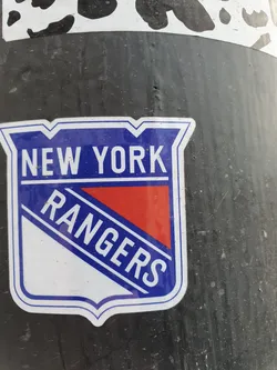A sticker featuring the New York Rangers logo. The logo is a shield shape with the words "NEW YORK" at the top and "RANGERS" at the bottom, in white lettering on a blue background. There's a red and white diagonal stripe in the middle.