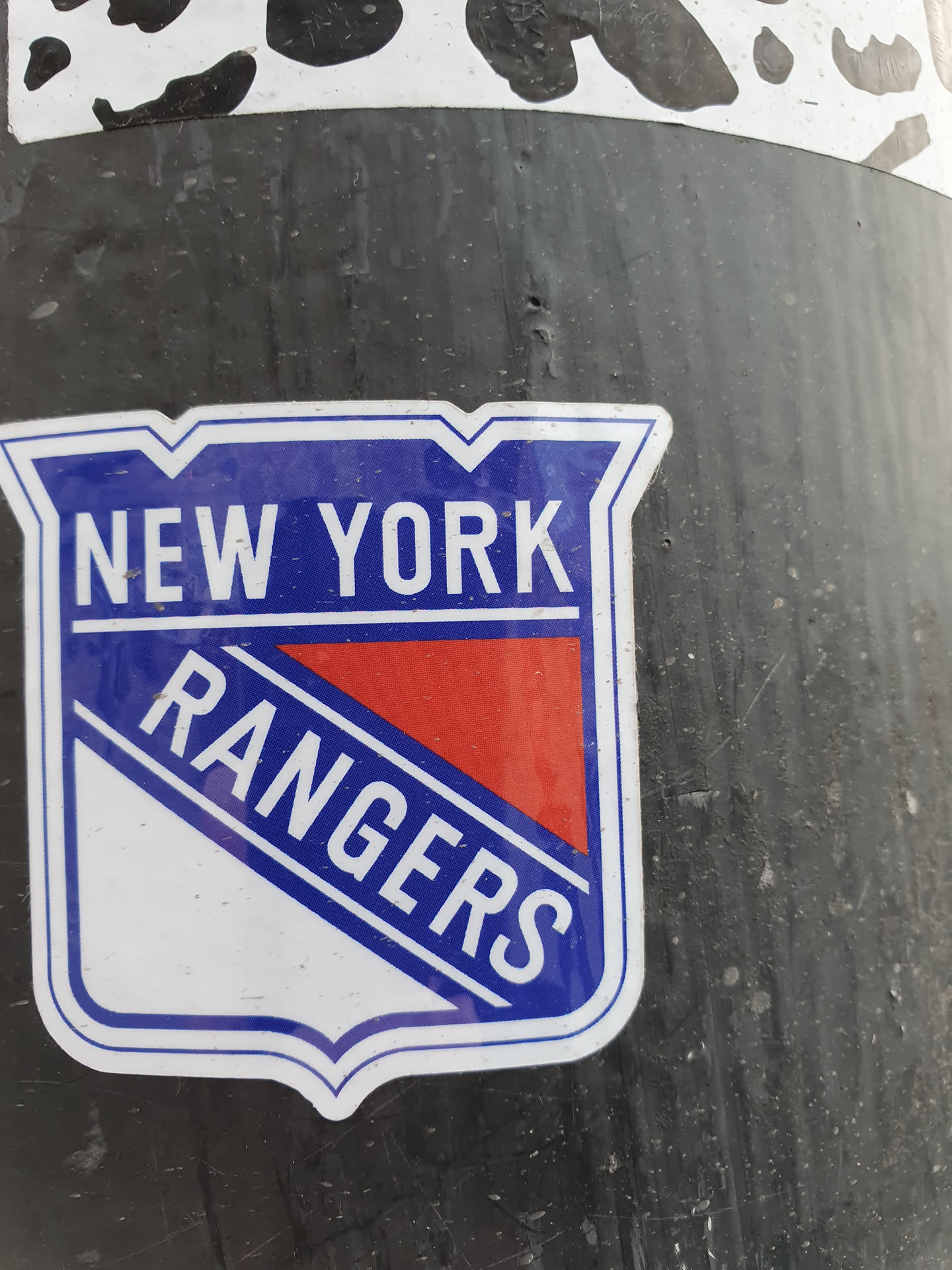A sticker featuring the New York Rangers logo. The logo is a shield shape with the words "NEW YORK" at the top and "RANGERS" at the bottom, in white lettering on a blue background. There's a red and white diagonal stripe in the middle.