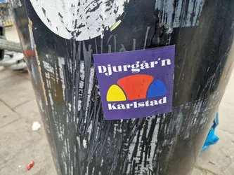 A purple sticker with the text "Djurgår'n Karlstad" and a stylized graphic of three overlapping semi-circles in yellow, red and blue.  The sticker is affixed to a dark-colored cylindrical surface covered in other stickers and markings.