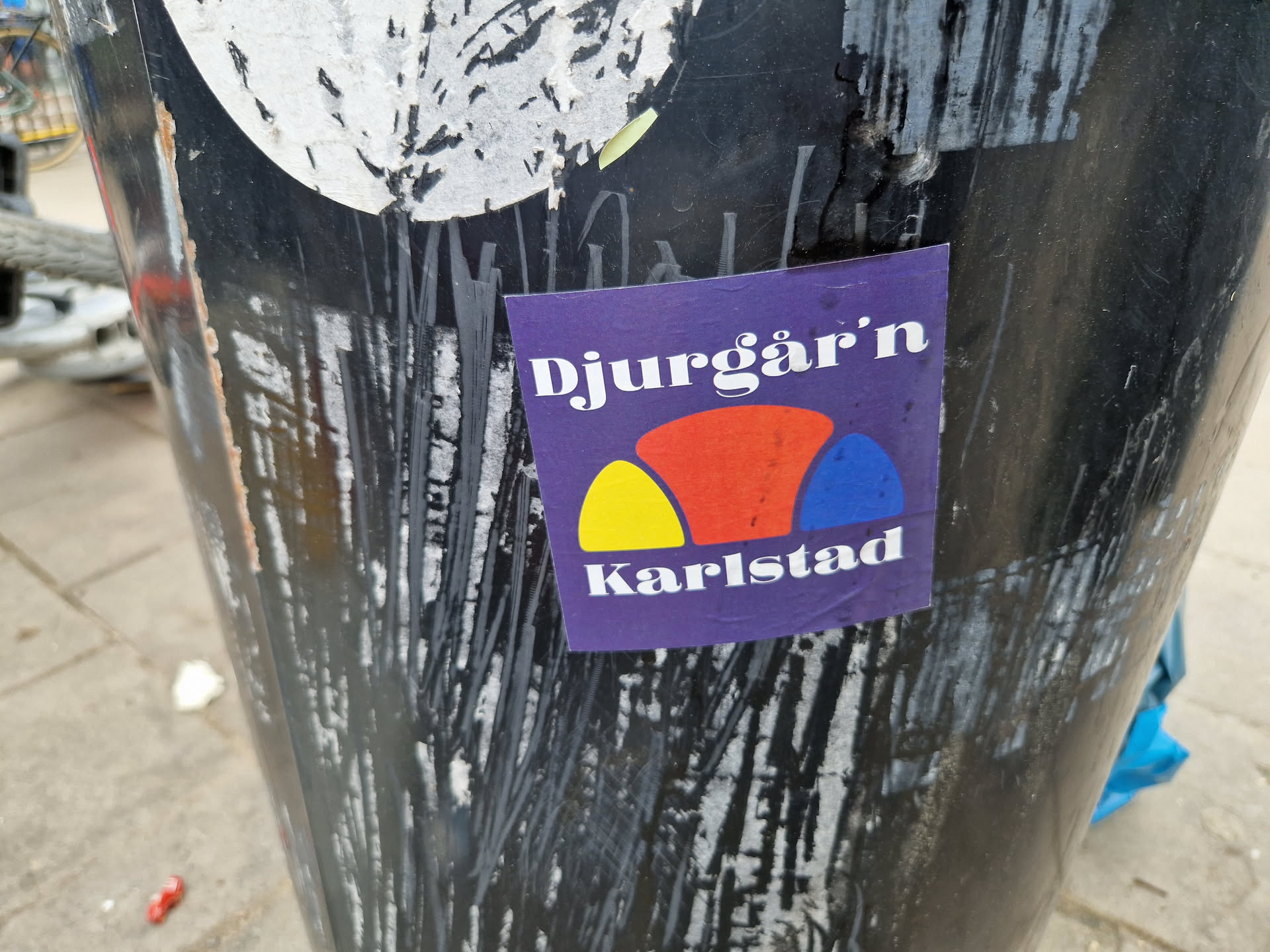 A purple sticker with the text "Djurgår'n Karlstad" and a stylized graphic of three overlapping semi-circles in yellow, red and blue.  The sticker is affixed to a dark-colored cylindrical surface covered in other stickers and markings.