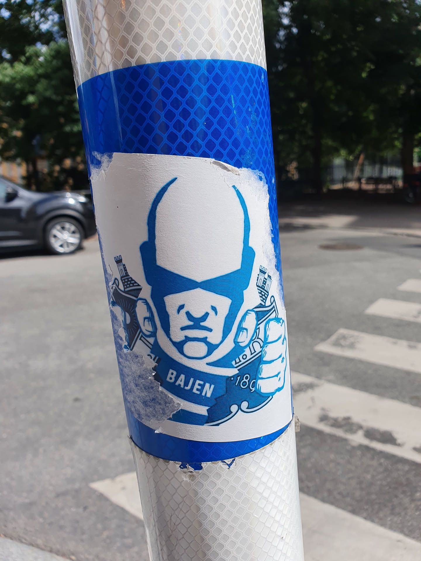 A blue and white sticker featuring a stylized face and the text "Bajen 1897".  The sticker is affixed to a reflective blue pole.