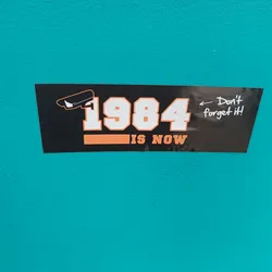 Black and orange sticker with the text "1984 IS NOW" and a picture of a security camera. It also has a handwritten note that says "Don't forget it!"