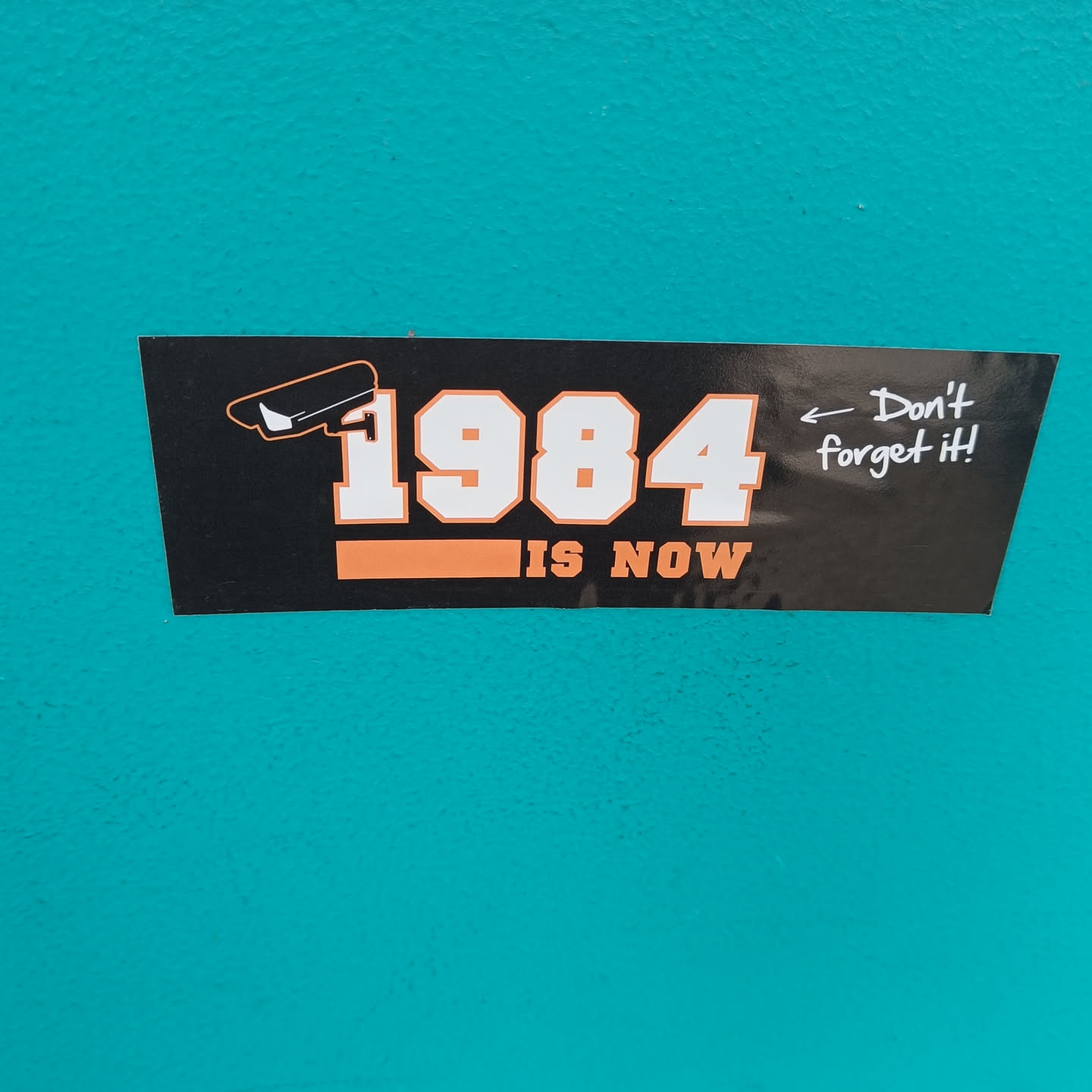 Black and orange sticker with the text "1984 IS NOW" and a picture of a security camera. It also has a handwritten note that says "Don't forget it!"