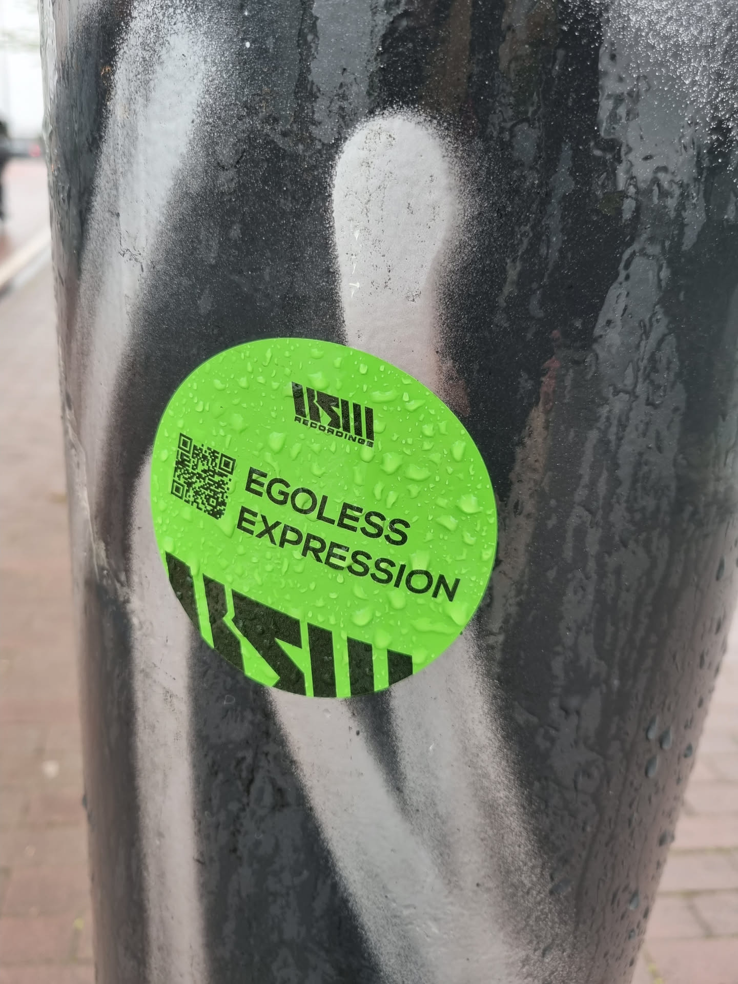 A round green sticker with a QR code and the text 'EGOless Expression'. The sticker has water droplets on it and is attached to a dark pole with white graffiti.  The logo 'KSII RECORDINGS' is also visible on the sticker.