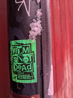 A rectangular, neon green sticker with bold black text and graphics, applied to a dark, possibly metallic pole. The sticker is slightly crumpled and shows signs of wear, with some snow visible on the pole above and to the right of it. The main text, 'FILM IS NOT DeAd', is arranged in a stacked, blocky, and somewhat distressed style. The 'O' in 'NOT' is replaced by a black smiley face with 'xx' eyes. Below this, in smaller black text, is 'STOCKHOLM INTERNATIONAL FILM FESTIVAL', accompanied by a small, stylized bird or plane icon. The green background has a subtle dotted or halftone texture.