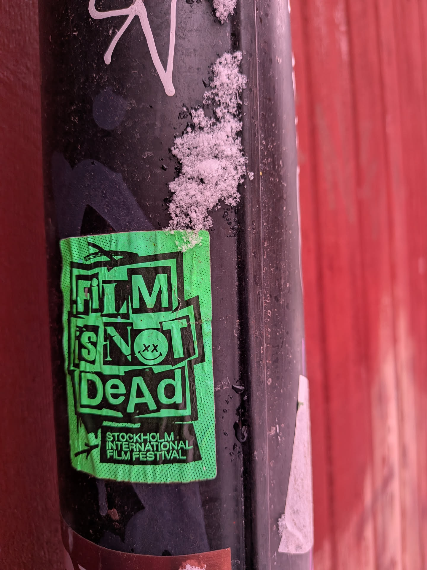 A rectangular, neon green sticker with bold black text and graphics, applied to a dark, possibly metallic pole. The sticker is slightly crumpled and shows signs of wear, with some snow visible on the pole above and to the right of it. The main text, 'FILM IS NOT DeAd', is arranged in a stacked, blocky, and somewhat distressed style. The 'O' in 'NOT' is replaced by a black smiley face with 'xx' eyes. Below this, in smaller black text, is 'STOCKHOLM INTERNATIONAL FILM FESTIVAL', accompanied by a small, stylized bird or plane icon. The green background has a subtle dotted or halftone texture.