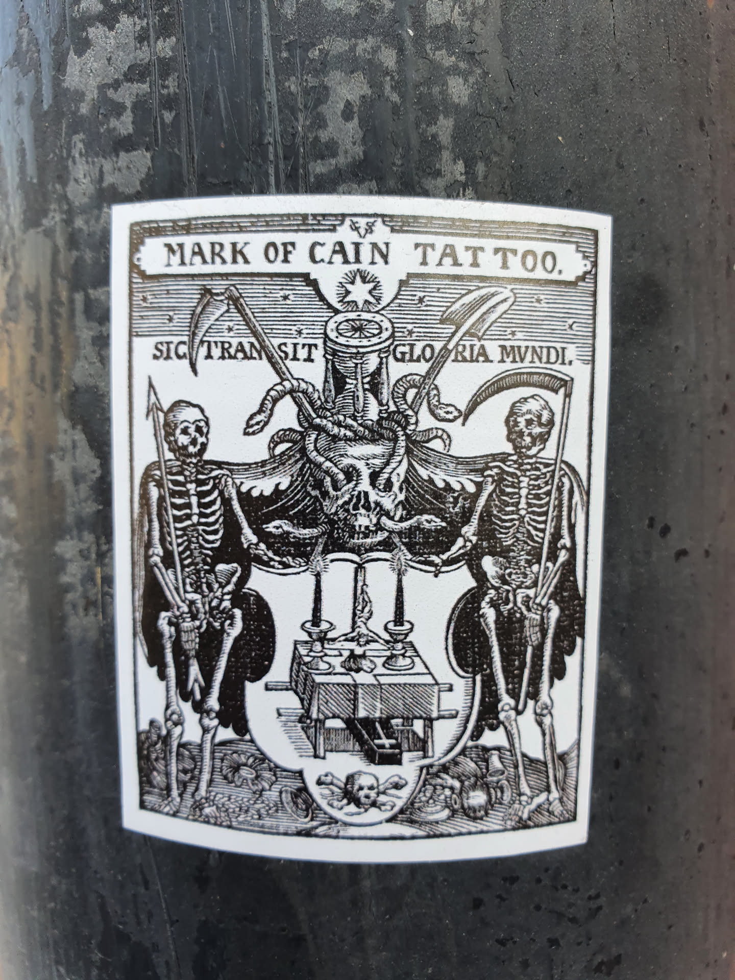Black and white sticker with a vintage woodcut style illustration featuring two skeletons, scythes, snakes, an hourglass, and a skull. The text at the top reads "MARK OF CAIN TATTOO."  Latin text is visible below the top text: "SIC TRANSIT GLORIA MUNDI". The sticker appears to be advertising a tattoo parlor.