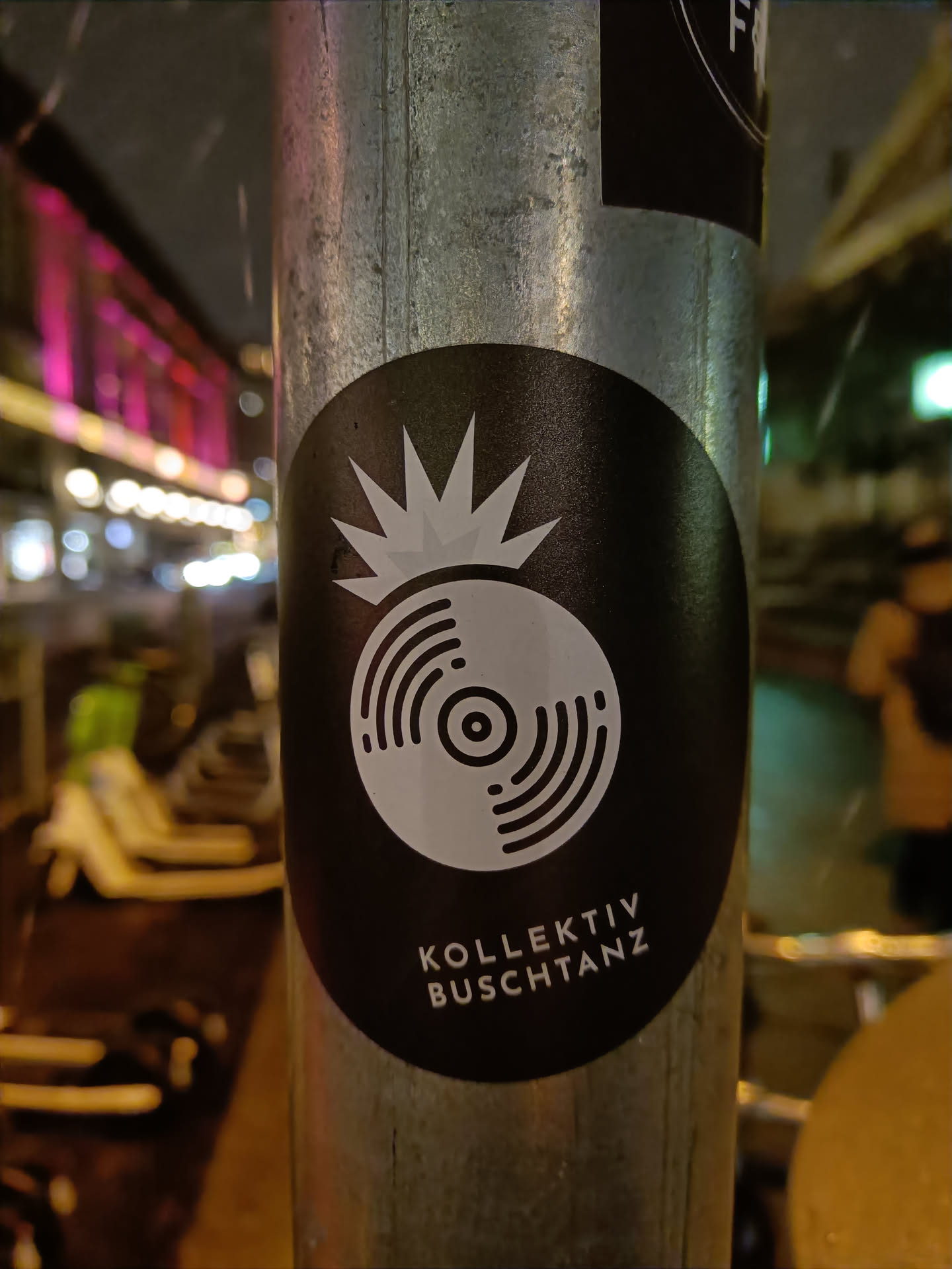 A black and white sticker with a vinyl record design and the text "KOLLKEKTIV BUSCHTANZ". The sticker is attached to a metal pole outdoors, suggesting it's street art. 