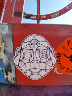 This sticker features a detailed black and white illustration of a face with geometric and abstract elements. The style is reminiscent of intricate line drawings and graphic design.  The face has large eyes and a prominent mustache, with various patterned details throughout. The sticker is affixed to a red metal surface amongst other stickers and graffiti.