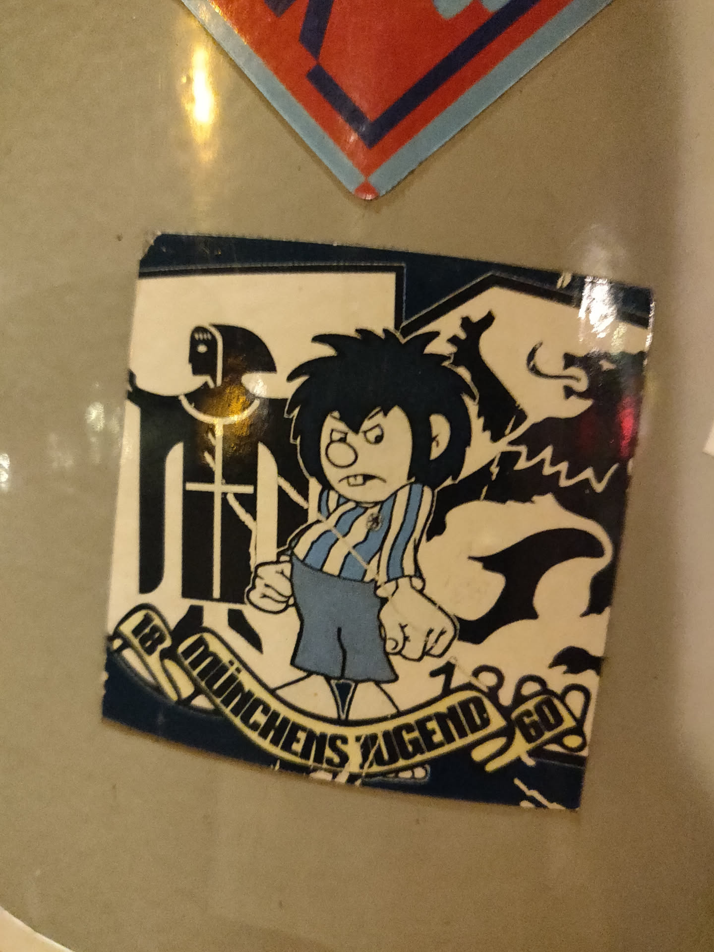 A sticker featuring a cartoon character with spiky hair, wearing a striped shirt and shorts. The character has an angry expression. The sticker also features a banner with the text "18 Münchens Jugend 60" and some other graphical elements.