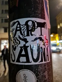 Black and white sticker with the words "Art Sauna" in a stylized font. The text is surrounded by abstract designs that look like smoke or steam.  The sticker is affixed to a pole outdoors at night.