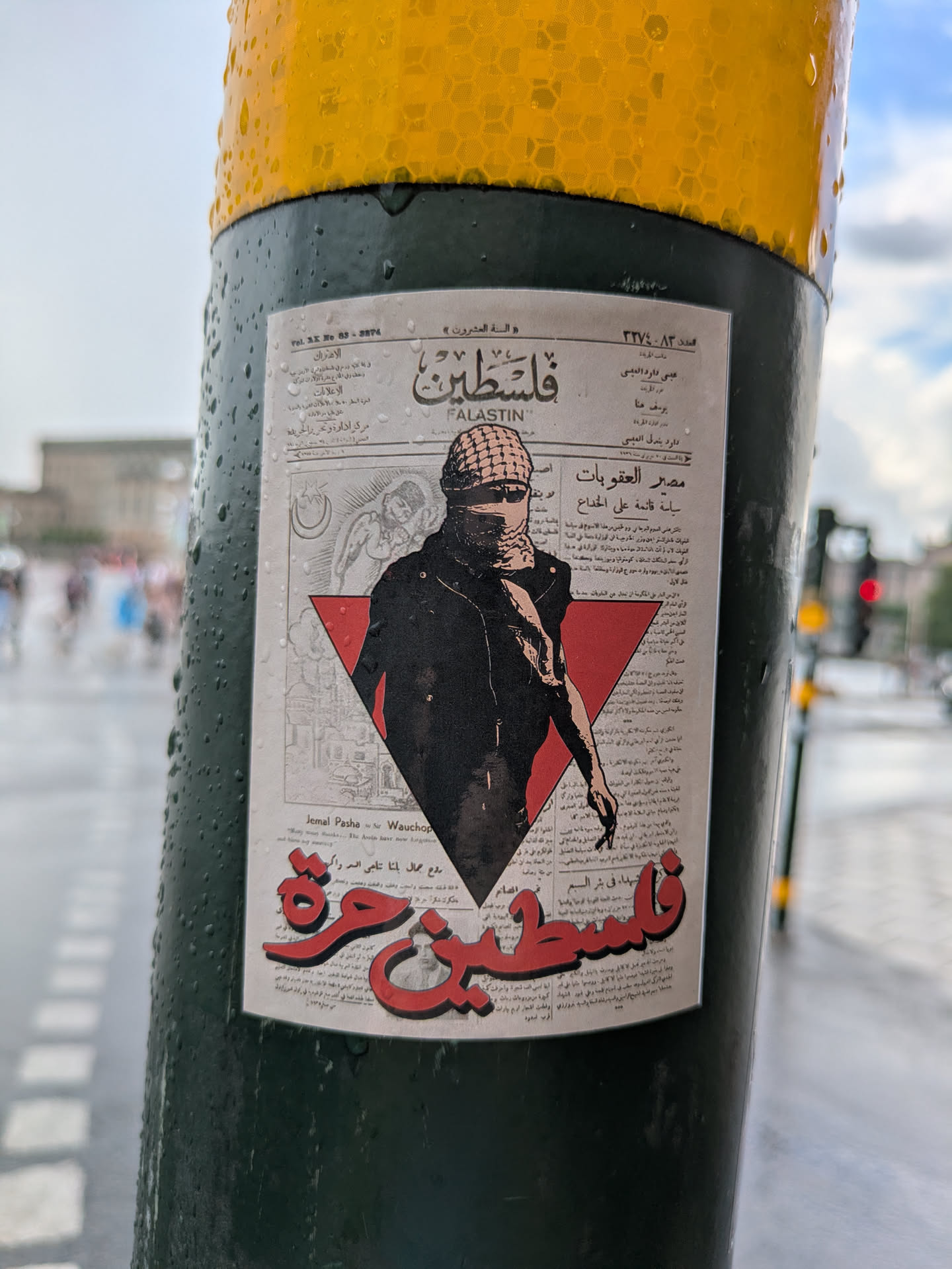 The sticker, designed to resemble an old newspaper clipping, features a central figure wearing a keffiyeh and sunglasses, with a determined expression. This figure is partially obscured by a prominent red inverted triangle, a symbol often associated with resistance. Behind the figure, faint grayscale illustrations depict historical or symbolic scenes, including a crescent moon and star, and what appears to be a building or landscape. The background is filled with small, dense Arabic text, mimicking newspaper articles. Large, stylized red Arabic text at the top reads 'فلسطين' (Falastin) and 'FALASTIN' in English, while equally prominent red Arabic text at the bottom declares 'فلسطين حرة' (Falastin Hurra), meaning 'Free Palestine'. The sticker is adhered to a dark green pole, which is visibly wet with raindrops, adding to its authentic street art appearance.